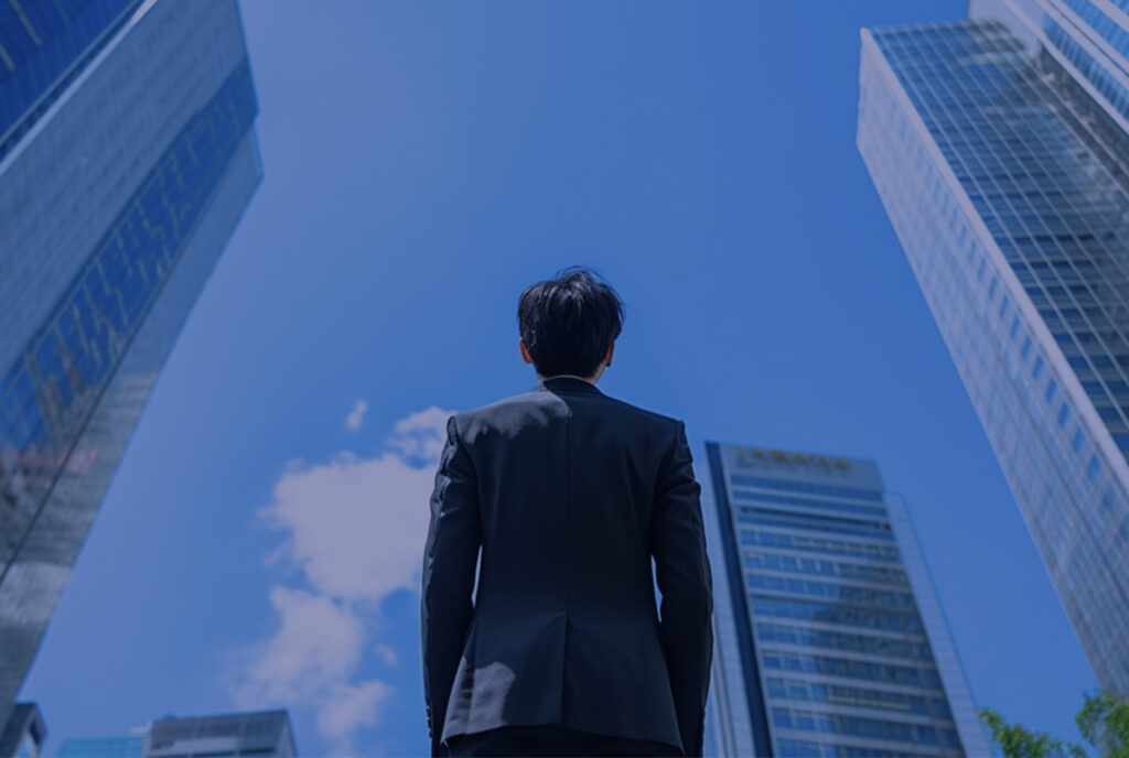 スーツを着た男性がビルの隙間に見える青空を仰ぎ見ている様子